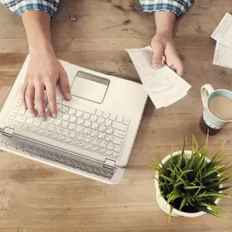 an open laptop, a hand holding a sheaf of bills, with more on the table, and a cup of coffee and a houseplant next to it